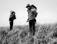 black and white photograph of three people standing in tall grass, one carrying a child on his back