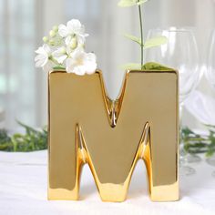 a gold m shaped vase with flowers in it sitting on a white table cloth next to wine glasses