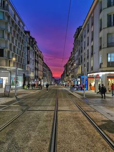 people are walking on the street in front of buildings at dusk, with train tracks running through the middle