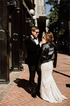 a bride and groom standing in front of a building on the street with their arms around each other