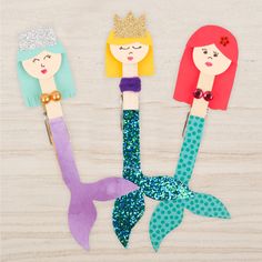 three little mermaids made out of popsicle sticks on a wooden table with glitter