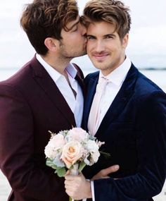 two men in suits are kissing each other on the beach while one man is holding a bouquet