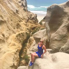 a woman in a blue swimsuit sitting on rocks near the ocean and looking at the camera