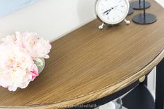 a wooden table topped with a vase filled with pink flowers next to an alarm clock