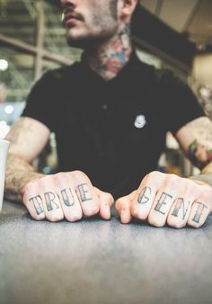 a tattooed man sitting at a table with two fingers crossed out and the word true bean written on his left hand