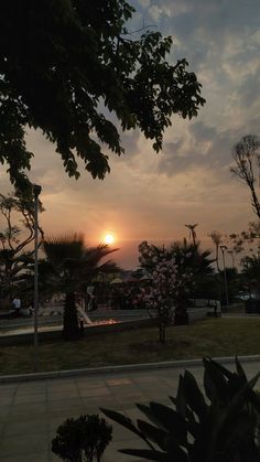 the sun is setting behind some trees and bushes in front of a park with people sitting on benches