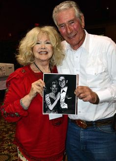 an older man and woman pose for a photo while holding up a black and white photograph