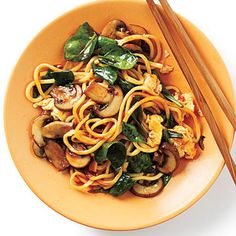 a yellow bowl filled with noodles and vegetables next to chopsticks on a white surface