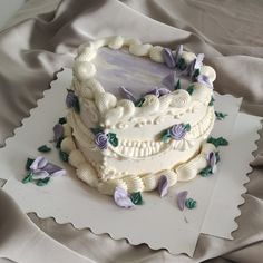 a white cake with purple flowers on it sitting on top of a cloth covered table