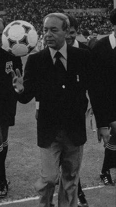 an older man holding a soccer ball in front of other men on the field at a stadium