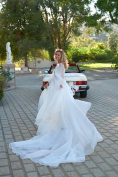 a woman in a white wedding dress standing next to a red car on a brick road