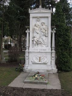 a monument with flowers around it in the middle of some grass and trees behind it