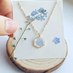 a hand holding a piece of jewelry next to a flower on a wooden slice with blue flowers in the background