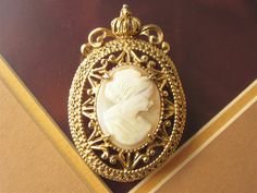 A beautiful signed Florenza cameo brooch, real carved shell portrait in an ornate goldtone setting with a crown at the very top. The backside has an attached bail if wanting to wear as a pendant by adding a necklace chain.  The brooch is 1 1/2 by 2 inch.  Mark:  Florenza In wonderful condition and ready to wear or give as a gift.  To see all the jewelry in my Etsy shop, go to my home page here ~ www.etsy.com/shop/darsjewelrybox Elegant Gold Cameo Brooch, Gold Cameo Brooch For Wedding, Antique Gold Cameo Brooch, Antique Gold Cameo Brooches, Formal Gold Cameo Brooches, Formal Gold Cameo Brooch, Gold Cameo Brooch Collectible, Gold Cameo Medallion Brooch, Gold Cameo Medallion Brooches