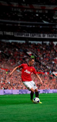 a man in red shirt kicking a soccer ball on green field with crowd watching behind him