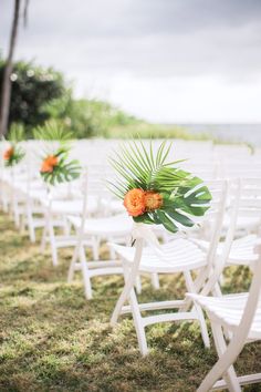 rows of white folding chairs with orange flowers and palm leaves on them in front of the ocean