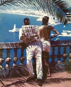 two people sitting on a bench looking out at the ocean with palm trees in the foreground