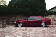 a maroon car parked in front of a stone wall and shrubbery on the other side