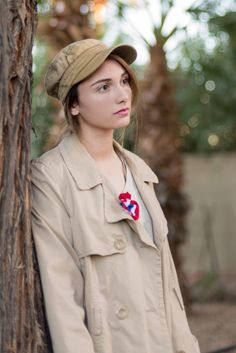 a woman standing next to a tree wearing a hat and trench coat with a flower on it