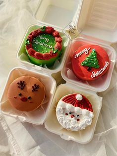 four different decorated cupcakes in plastic containers on a white tablecloth with writing merry christmas