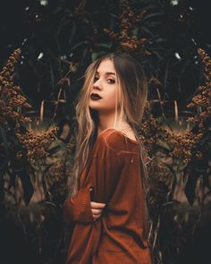 a woman with long hair standing in front of some plants and looking into the distance