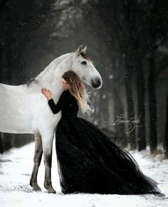 a woman in a black dress standing next to a white horse on a snowy day