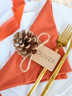 an orange napkin with a pine cone on it next to a gold fork and knife