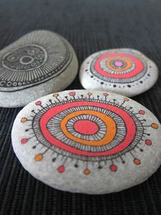 three rocks with designs painted on them sitting on a black tableclothed surface and one has an orange circle in the middle