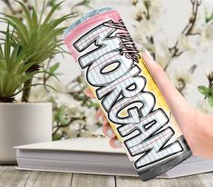 a hand holding a can of energy drink on top of a wooden table next to a potted plant