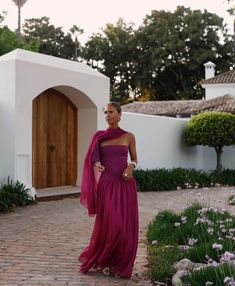 a woman in a purple dress is standing on a brick walkway with flowers and bushes around her