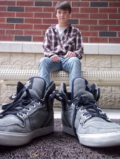 a young man sitting on top of a pair of gray high - top sneakers in front of a brick wall