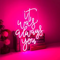 a pink neon sign that says it was always you next to a stack of books