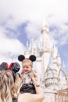 a woman holding a baby in front of a castle with mickey mouse ears on it