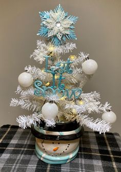 a small white christmas tree with snowflakes and ornaments in a pot on a plaid tablecloth