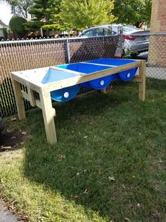 a wooden table with two blue plastic barrels on it in the grass next to a chain link fence