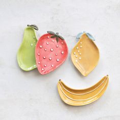 three ceramic fruit shaped like faces on a white surface, one with a green banana and one with a red strawberry