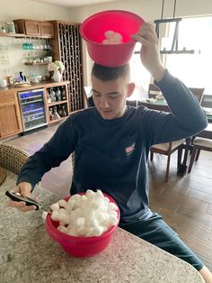 a man sitting at a table with a red bowl on his head and marshmallows in it