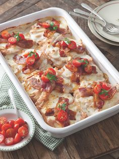 a casserole dish with tomatoes and meat in it on a wooden table next to silverware