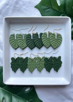 four green leaf shaped earrings on a white plate next to some leaves and greenery