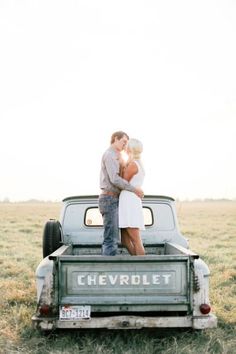 a couple standing on the back of an old pickup truck in a field with their arms around each other