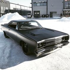 an old black car parked in the snow near a building and parking lot full of snow