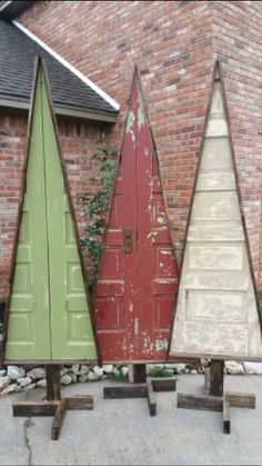 three wooden christmas trees sitting in front of a brick building with red doors and green shutters