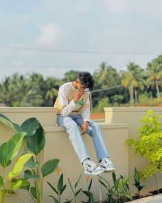 a man sitting on top of a cement wall next to plants and talking on a cell phone