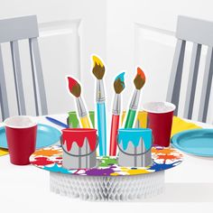 there are paintbrushes and cups on the table with paper plates in front of them