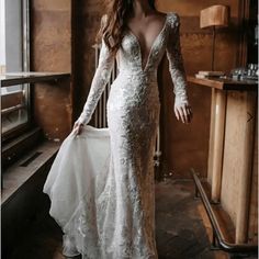 a woman in a long sleeved wedding dress standing by a window with her hand on her hip