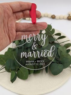 a hand holding a merry and married ornament on top of a plate with greenery