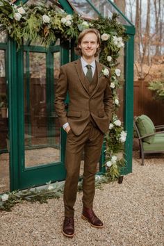 a man in a brown suit and tie standing next to a green glass building with flowers on it