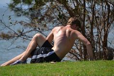 a shirtless man laying on the grass next to a tree and body of water