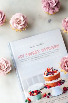 a cookbook with pink frosted cupcakes and flowers on the table next to it