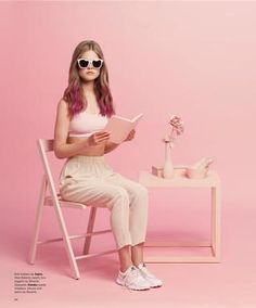 a woman sitting in a chair reading a book with sunglasses on her head and pink background